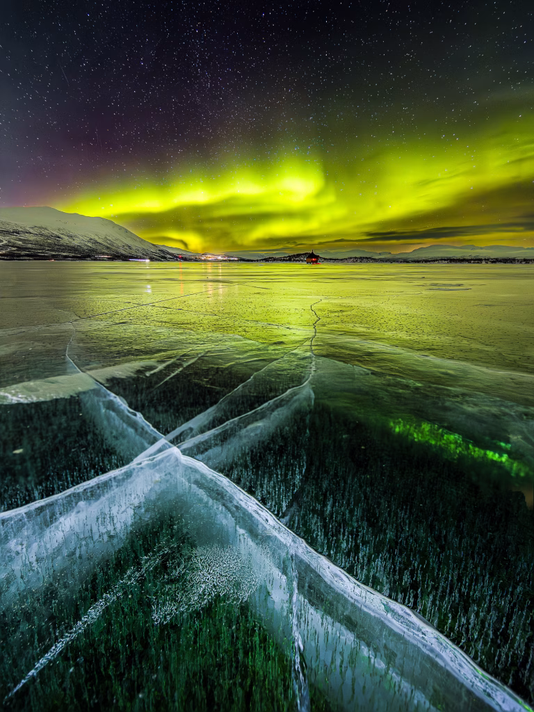 Clear ice with cracks and trapped air bubbles on Lake Torneträsk reflecting northern lights near Abisko with Mount Noulja and an ice fishing hut in the distance